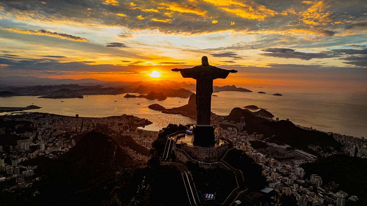 The statue and summit silhouette against the setting sun, with the city, bay, and layered mountain peaks below in Rio de Janeiro, Brazil.